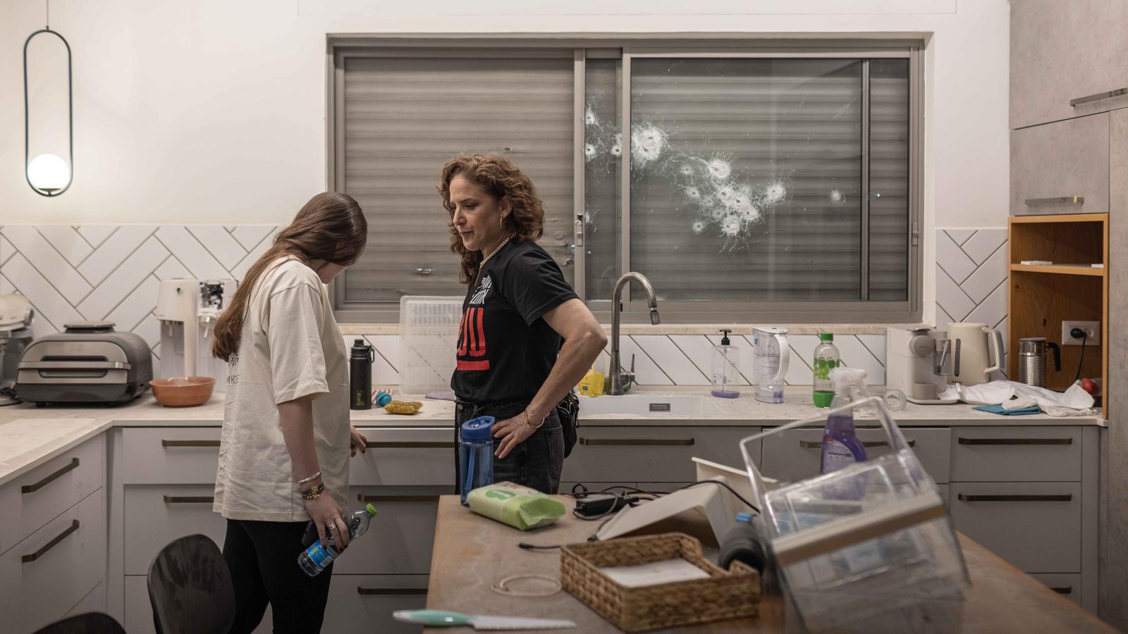 A woman and teenager in a messy kitchen. There are bullet holes in the window behind them.