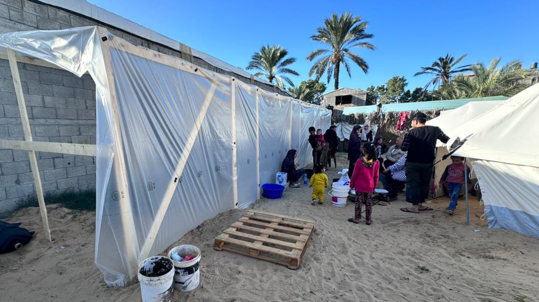 A group of people, including some children, stands in the distance amidst some tents and tarp.